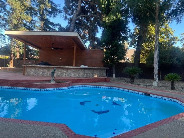 Curved blue swimming pool with brick edging, modern wooden pool house, and tall trees in background under clear sky