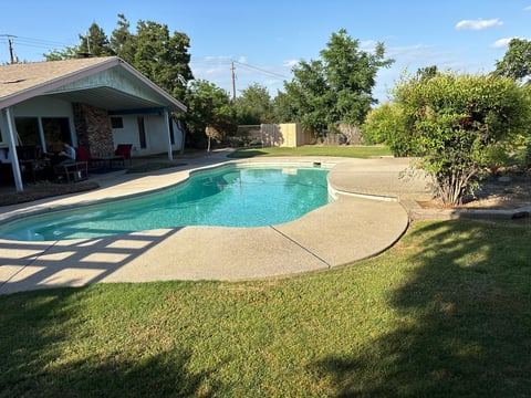 Backyard with turquoise swimming pool, concrete deck, green lawn, pool house with covered patio, and surrounding trees on sunny day