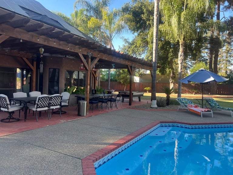 A backyard pool area with a covered wooden patio, lounge chairs, and trees on a sunny day