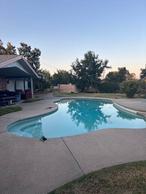 Backyard swimming pool with turquoise water and concrete deck, bordered by lawn and trees at dusk